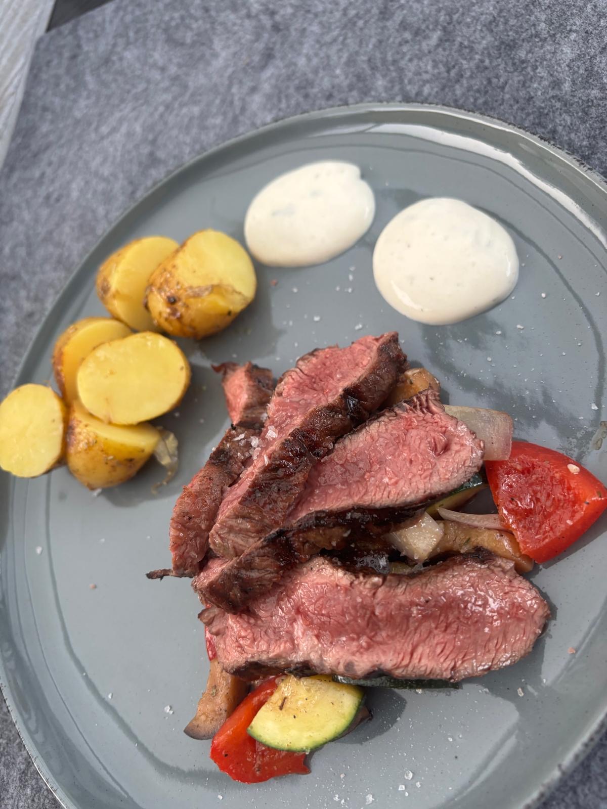 Saftiges Rindersteak in Scheiben mit Bratkartoffeln und cremiger Sauce, serviert mit Tomate auf grauem Teller, herzhaftes Hauptgericht