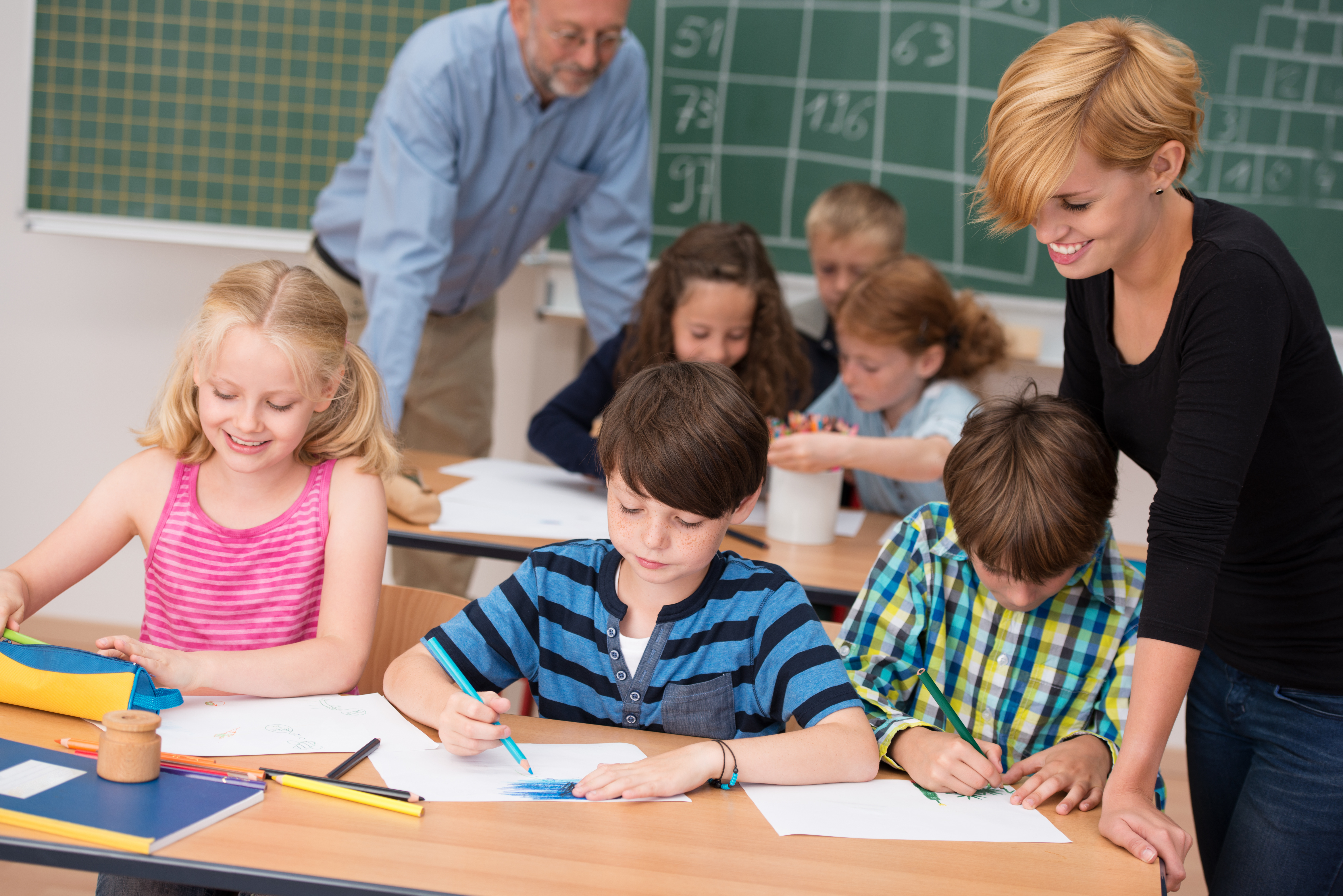 Pädagogische Mitarbeiterin unterstützt Kinder beim Lernen im Klassenzimmer, Grundschüler arbeiten konzentriert an Aufgaben mit Heften und Stiften, Förderung von Bildung und Konzentration