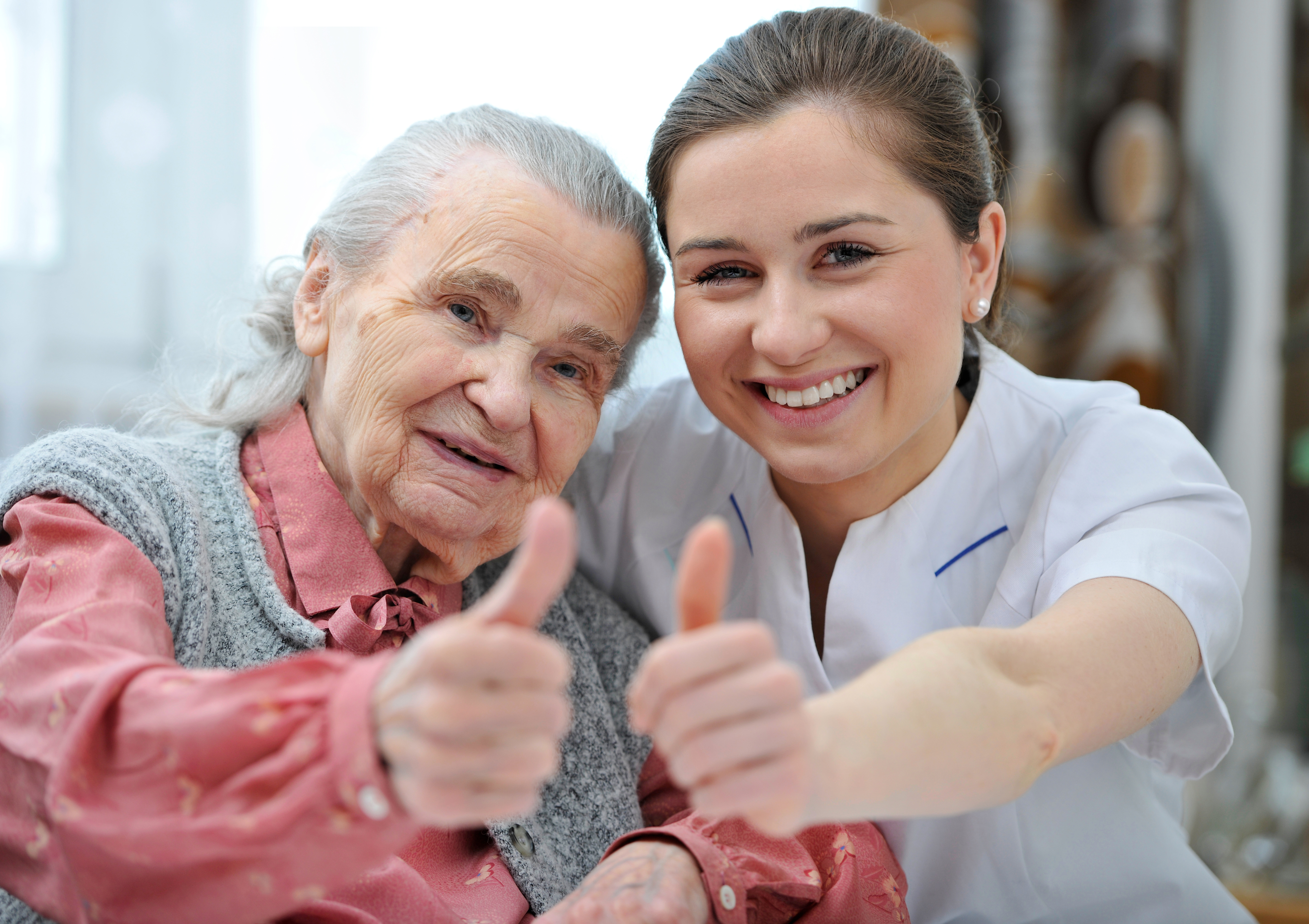 Pflegekraft und Seniorin zeigen Daumen hoch, positive Betreuung und Zufriedenheit in der Altenpflege, freundliche Unterstützung im Pflegealltag